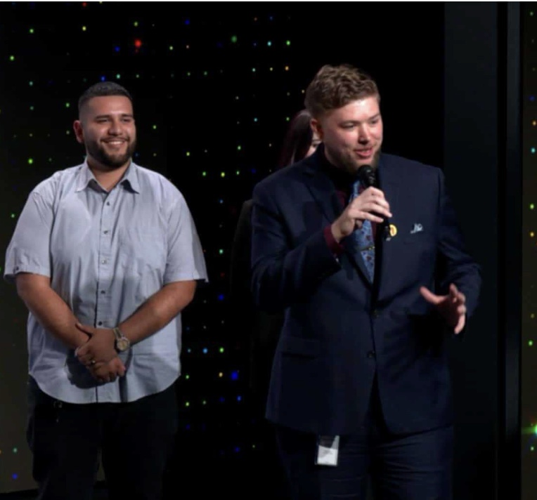 Marcus Gates in a navy suit speaking into a microphone on the Snap Design Academy showcase stage, with a teammate standing beside him